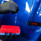 Ceramic coating being dropped onto a red microfibre applicator beside a blue Subaru WRX