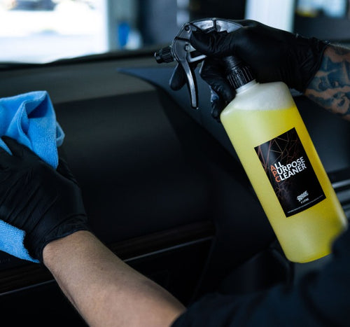 Gloved hand wiping a car dashboard with a microfibre cloth while holding Deluxe Detail All Purpose Cleaner